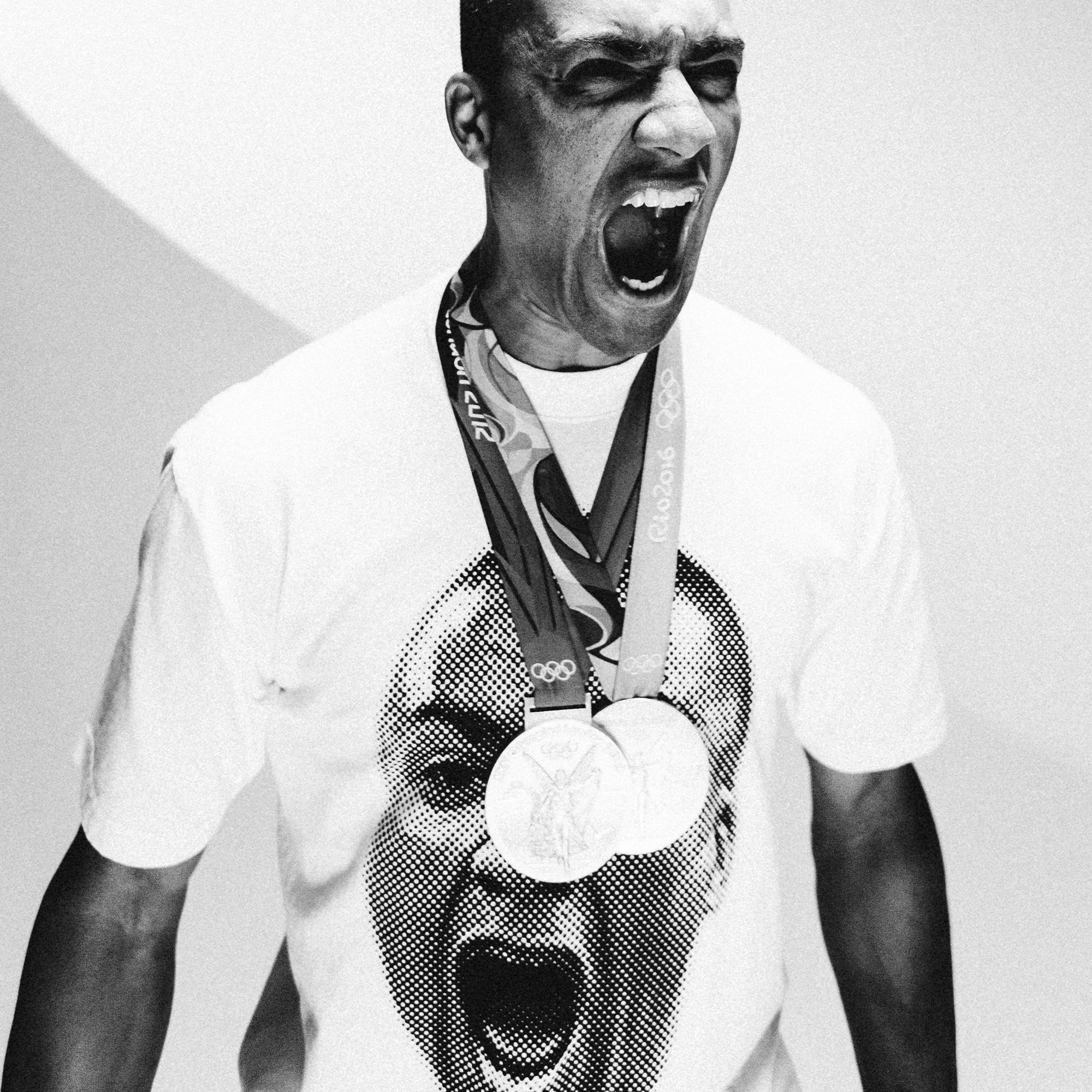 A man adorned with several Olympic medals passionately shouts in celebration, wearing the All-American Ashton Eaton Oversized Tee, which features a large, printed image of his own shouting face. The simple and unobtrusive background highlights his emotional expression as one of the greatest athletes of our time.