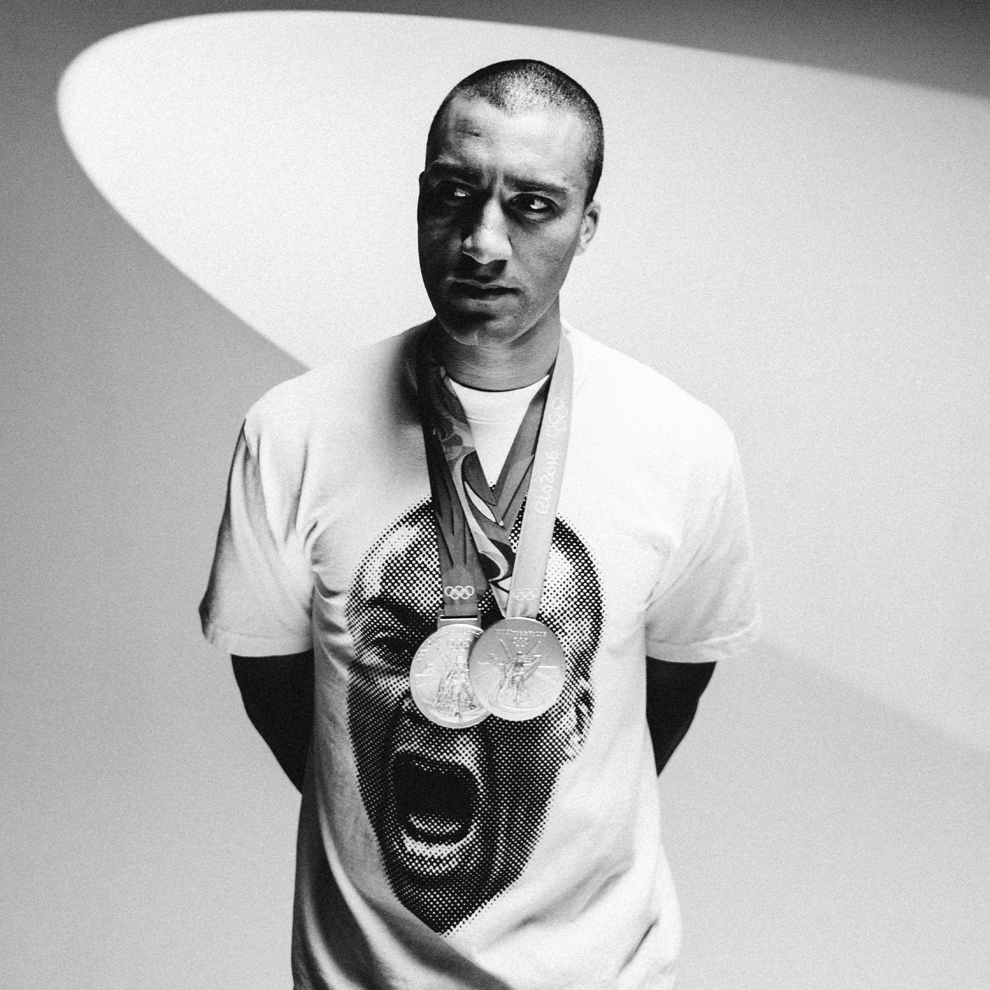 A person with a shaved head, wearing the All-American Ashton Eaton Oversized Tee that features a stylized face, stands against a minimalist backdrop. They have multiple medals hanging around their neck, exuding the aura of the greatest athlete. The image is in black and white, capturing a 2024 Olympic-inspired moment as they look slightly to the side with a serious expression.