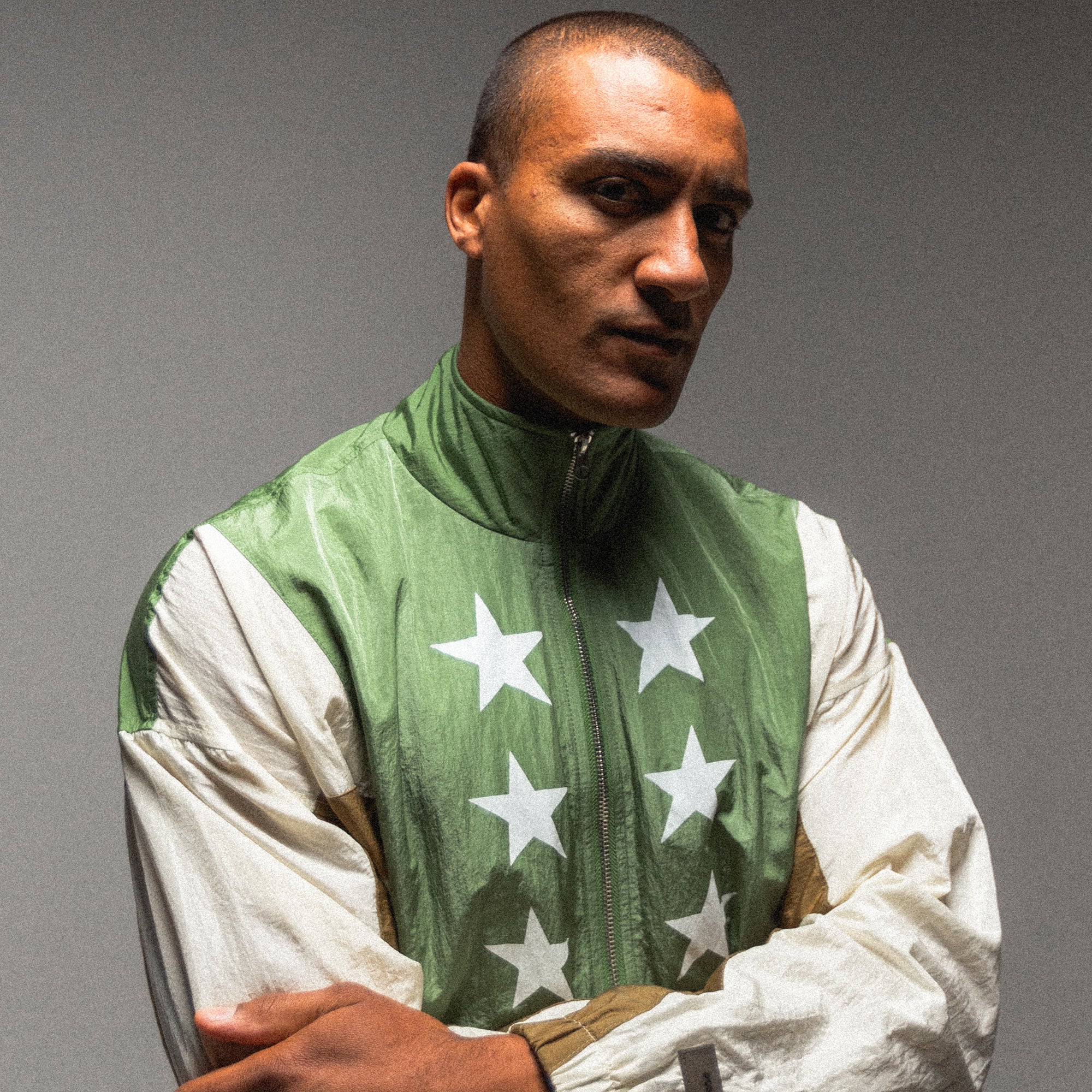 A person with a serious expression stands with arms crossed, donning the 2024 Olympic-inspired Olympic Summit Track Jacket in green and white, featuring white stars on the chest and sleeves. The plain gray background evokes the focused intensity of an athlete like Ashton Eaton.