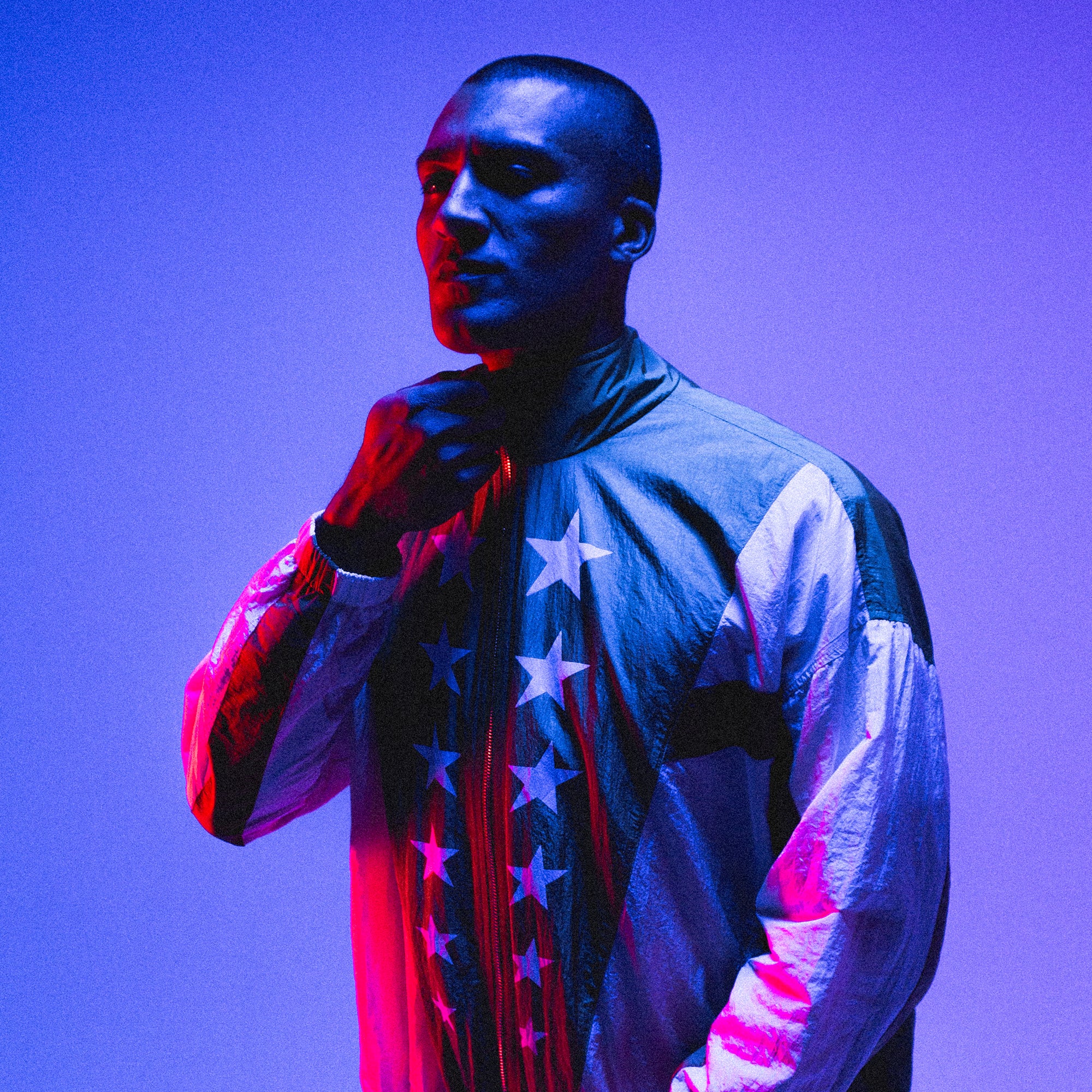 A person stands against a vibrant blue and purple background, adjusting the collar of an Olympic Summit Track Jacket adorned with stars. The lighting creates strong shadows, emphasizing the silhouette and features of the individual.