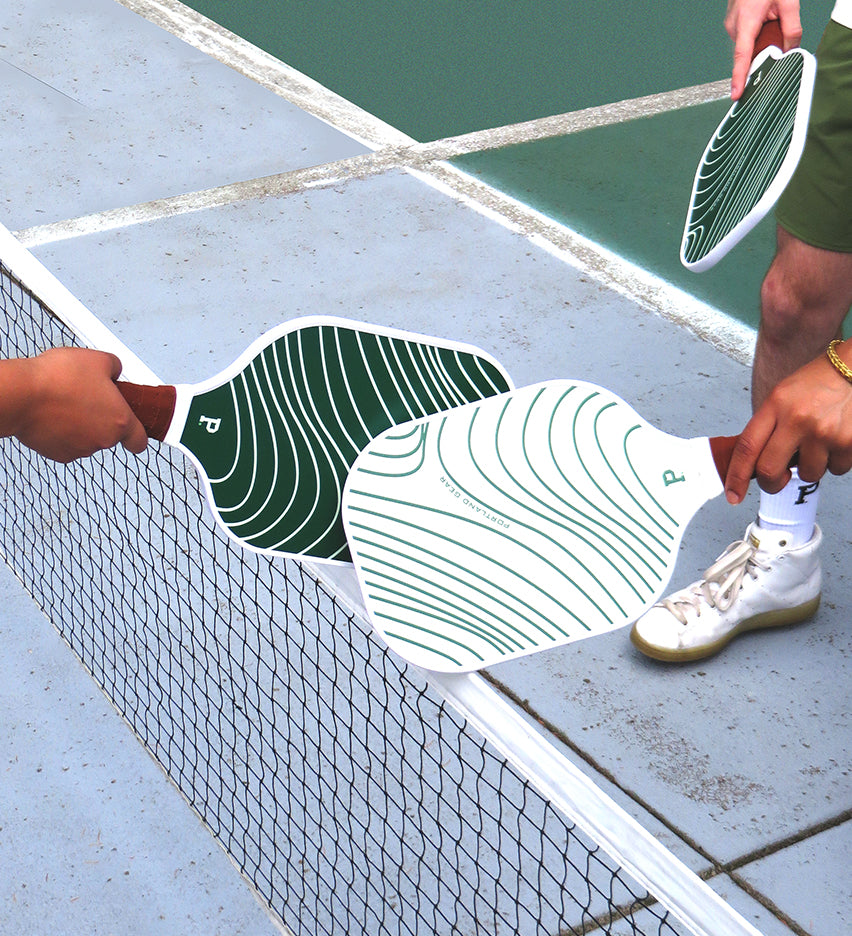 Close-up of three individuals standing on a pickleball court, each holding a limited edition Pickleball Paddle from Portland Gear and reaching their paddles over the net to tap them together in a pre-game gesture. The paddles feature a green and white wavy pattern.