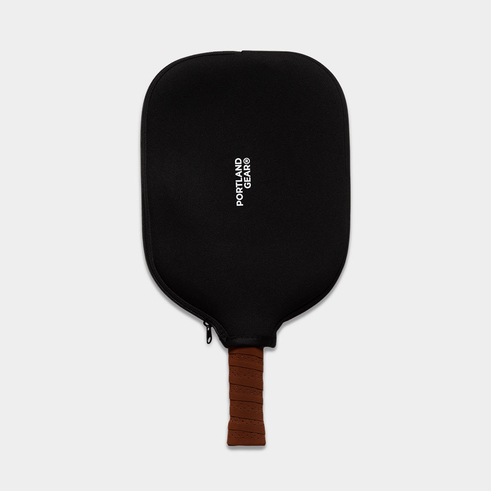 A black pickleball paddle cover features "PORTLAND GEAR" printed in white text vertically. Partially visible below the cover is a limited edition Pickleball Paddle, revealing a brown, textured handle indicating it is stored inside the cover. The background is a plain light gray.