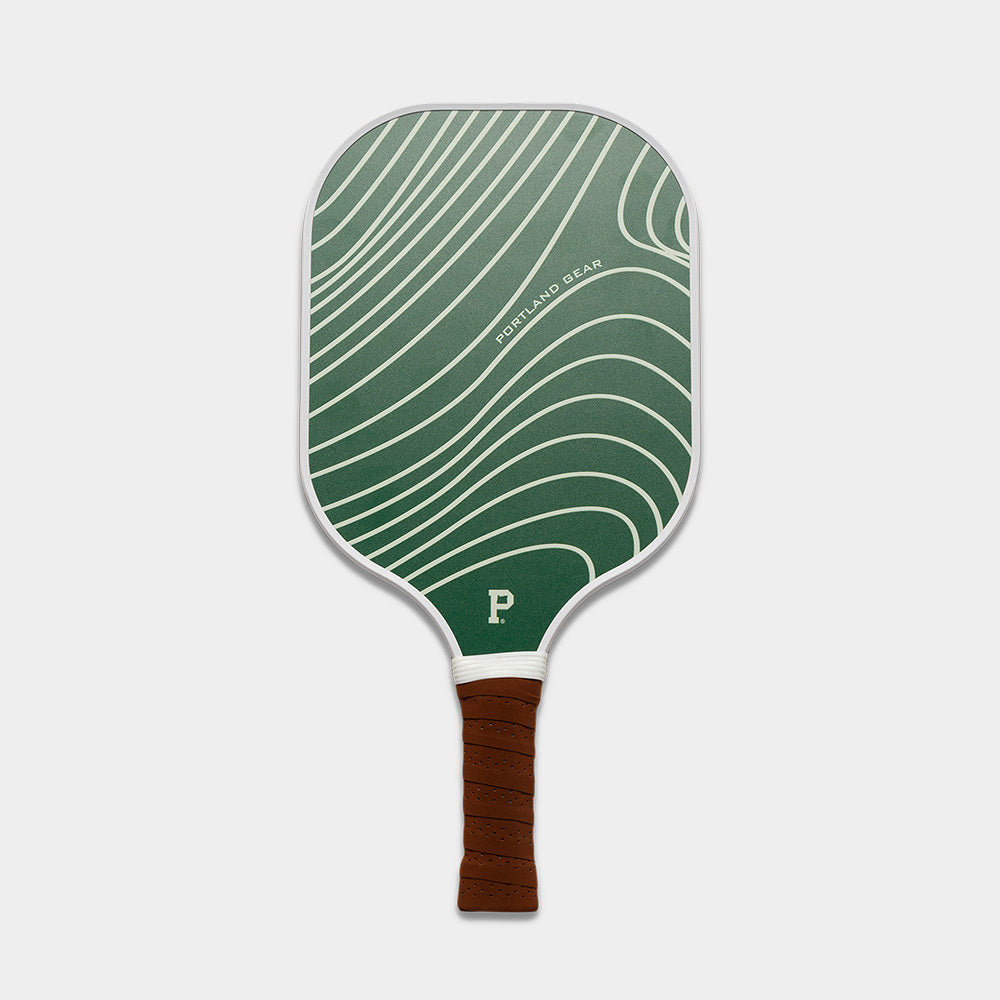 Introducing the limited edition Pickleball Paddle by Dreamland Gear, featuring a striking green face adorned with wave-like white lines and the brand name prominently displayed. The handle is wrapped in premium brown grip tape for optimal comfort, and the base of the paddle face showcases a distinctive white "P" logo inspired by Portland Gear. All set against a light grey background, this paddle is both functional and visually appealing.