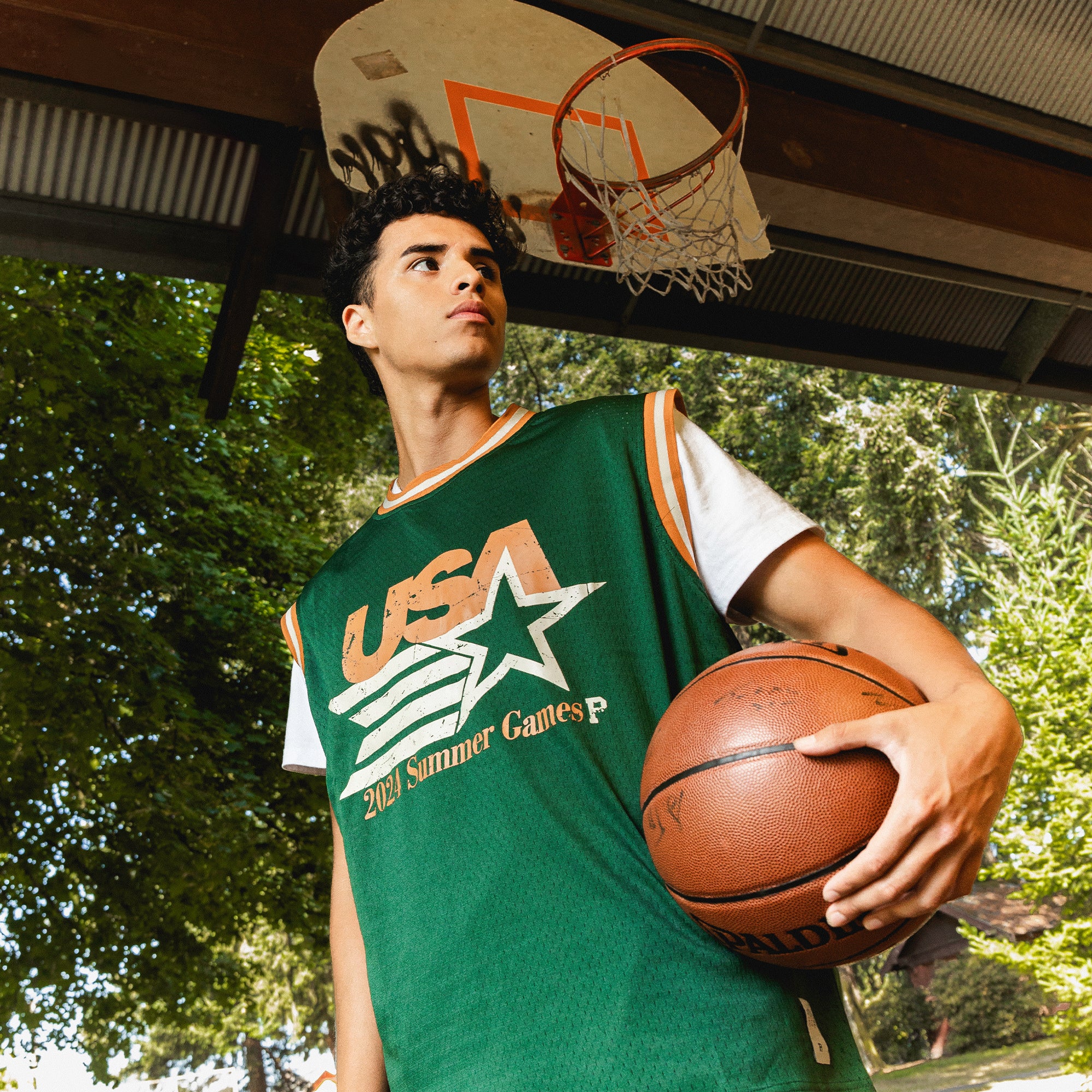 A young man stands outdoors beneath a basketball hoop, wearing a green "Olympic Mesh Jersey" over a white T-shirt. He holds a Spalding basketball under one arm and looks off into the distance, with lush green foliage in the background.