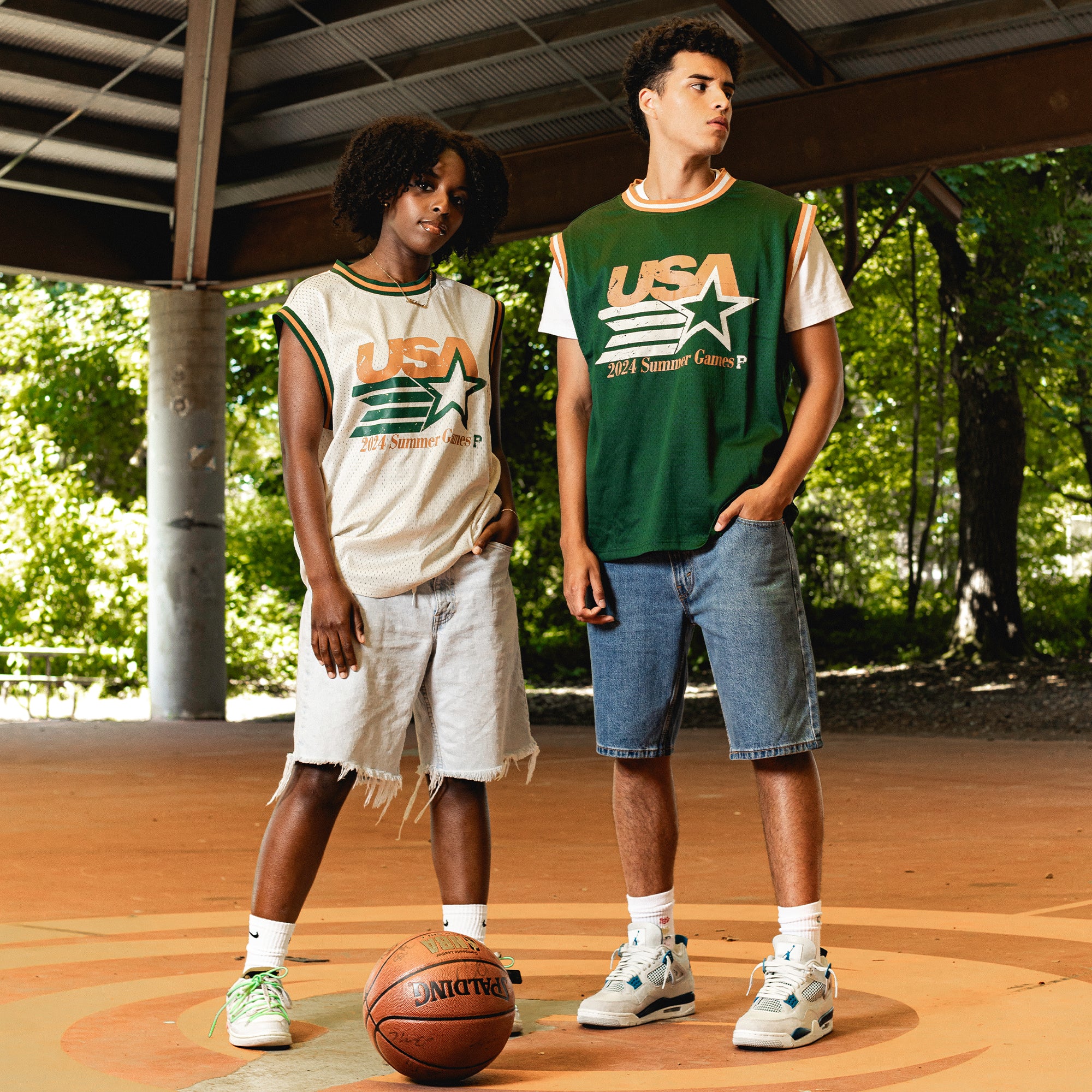 Two people, both wearing Olympic Mesh Jerseys with a USA theme, stand on a basketball court under a large shelter. The individual on the left is dressed in a white Olympic Mesh Jersey accompanied by ripped shorts, while the one on the right sports a green Olympic Mesh Jersey and denim shorts. A basketball lies on the ground nearby, suggesting their 2024 Olympics-inspired practice.